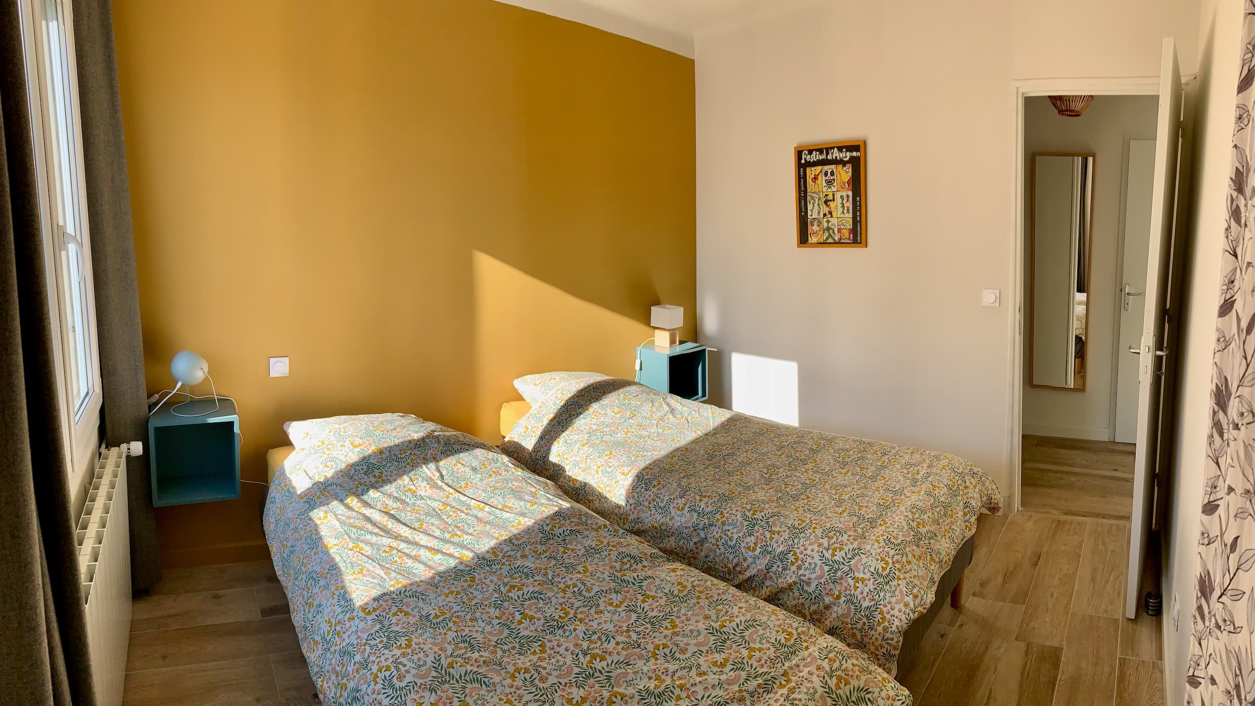 Vue de la chambre en soirée avec les deux lits, le mur jaune, l'affiche, la porte, et les lampes de chevet éclairées