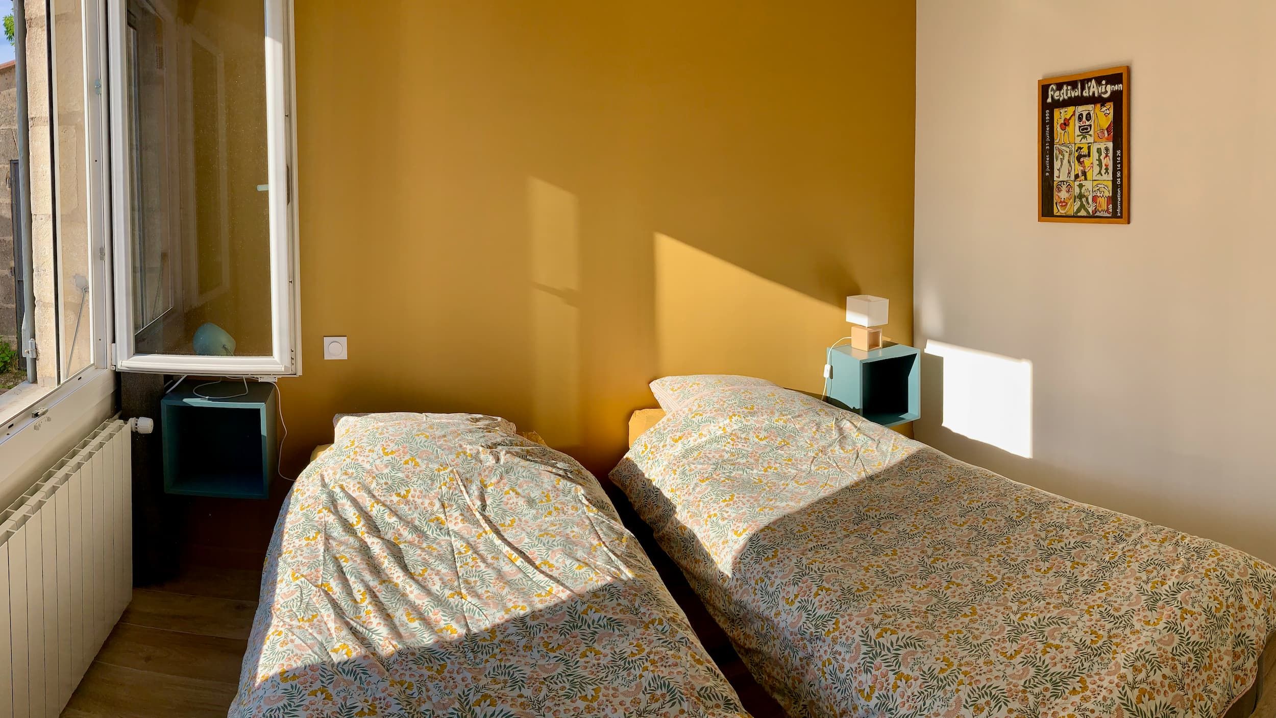 Vue de la chambre en soirée avec les deux lits, le mur jaune, l'affiche, la porte, et les lampes de chevet éclairées