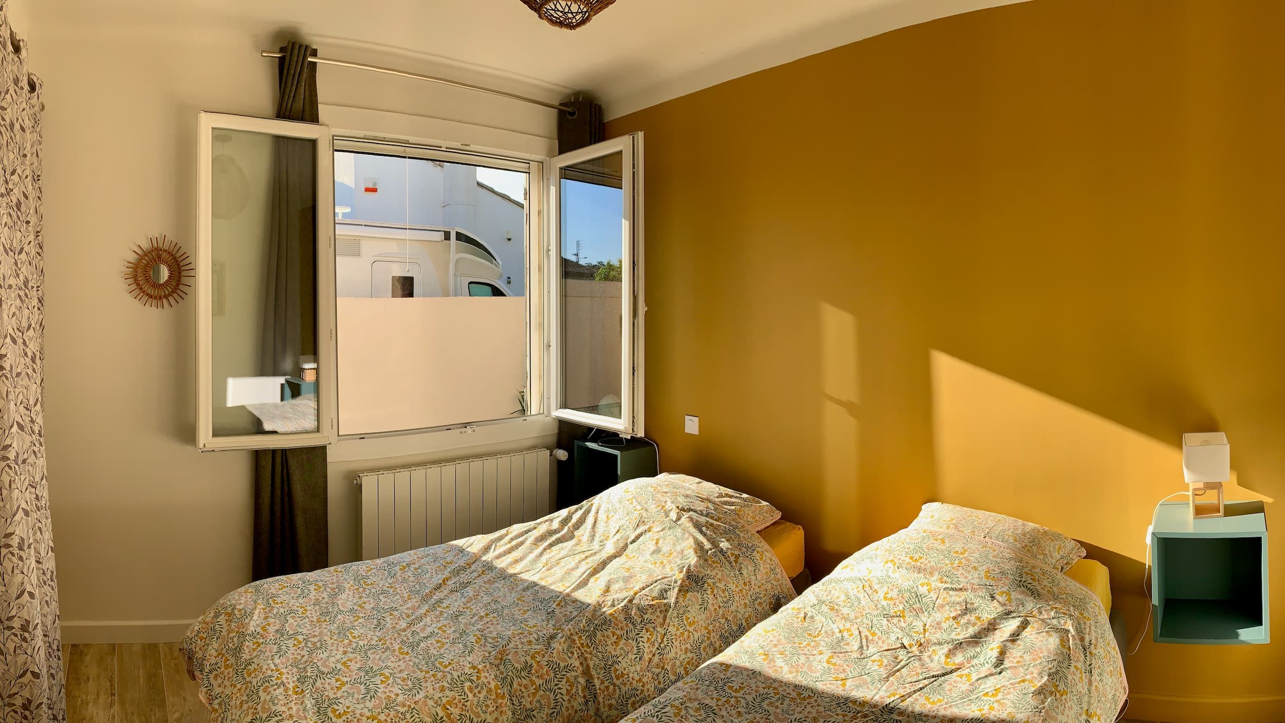 Vue de la chambre en journée avec les deux lits, le mur jaune, l'affiche, et les lampes de chevet