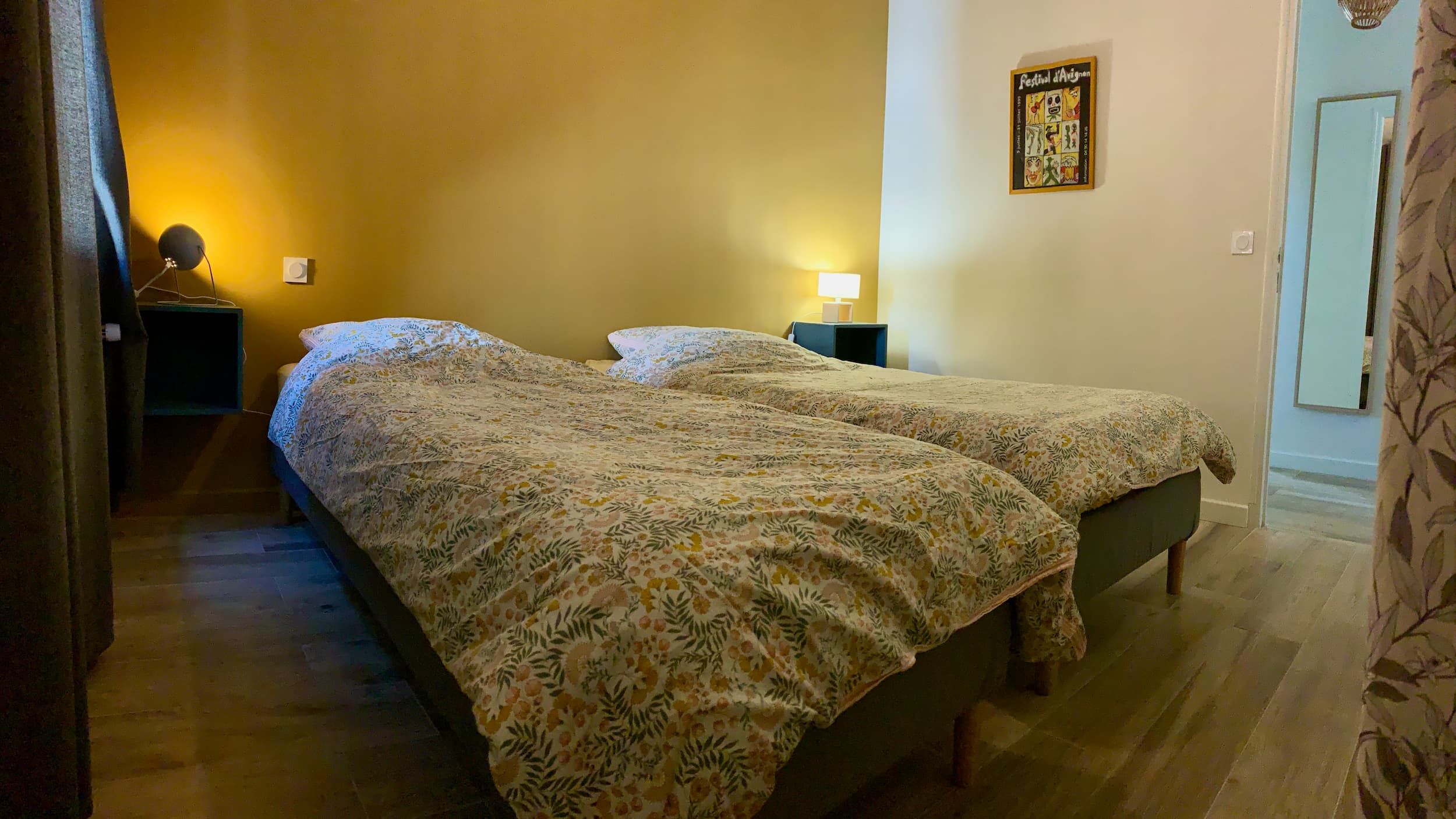 Vue de la chambre en journée avec les deux lits, le mur jaune, le mirroir, la fenêtre, et les lampes de chevet